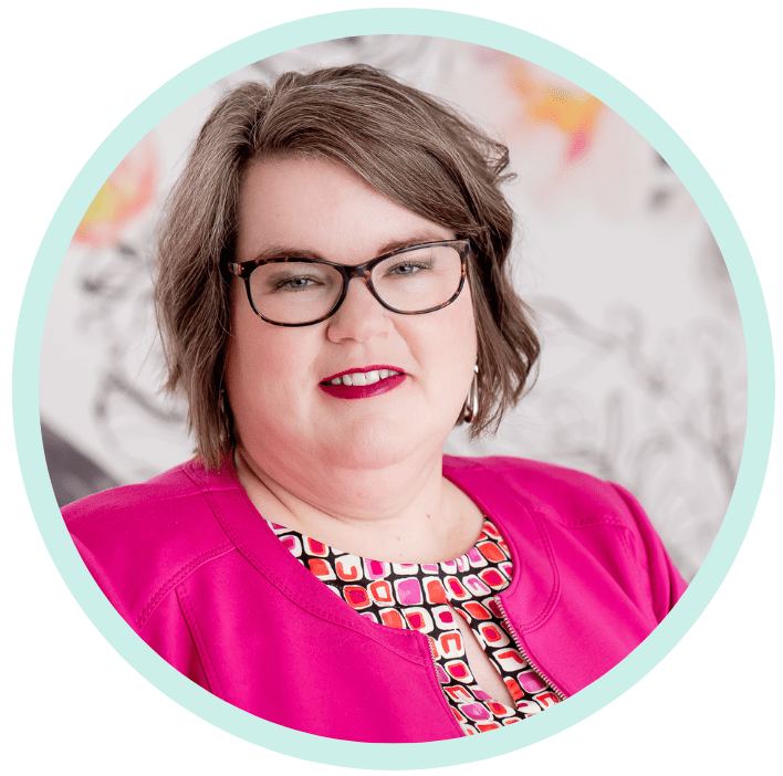 Kelly is a white woman, wearing glasses and a pink dress jacket, standing against a floral wallpaper background.