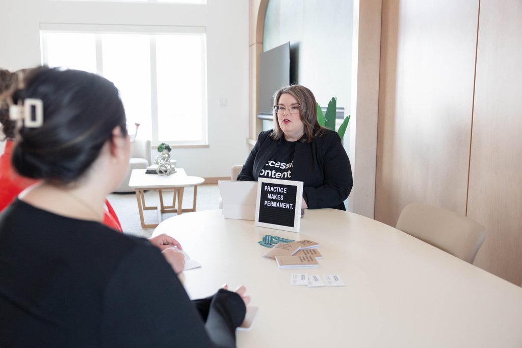 Kelly sits across a table from two people. She's wearing a black jacket and t-shirt with a sign showing the words practice makes permanent.